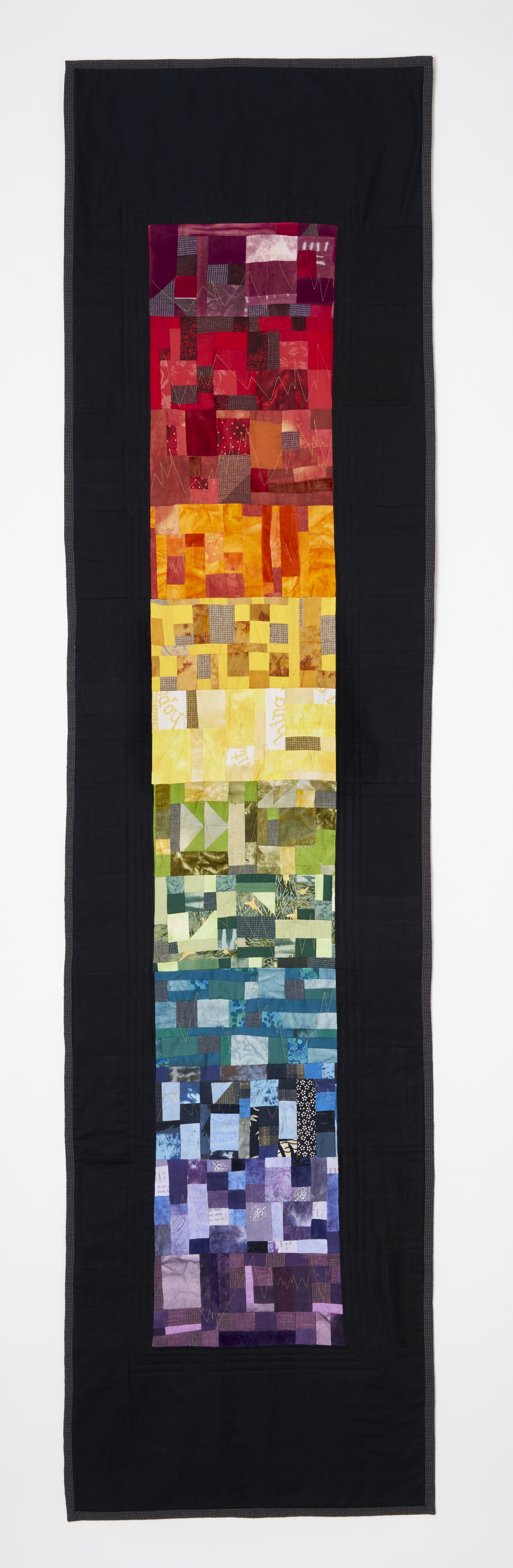 long narrow quilt with pieced colored rectangles that go through twelve colors of the rainbow from redviolet to violet and gold zigzag stitching